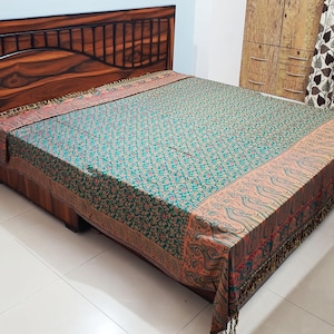 May include: A rectangular bedspread featuring a dark teal centre with a dense, repeating floral pattern in gold and red. The border displays a prominent paisley motif in shades of orange, red, and green, finished with decorative fringe along the edges.