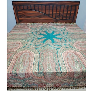 Puede incluir: Una colcha con un intrincado estampado de cachemira en tonos turquesa, rosa y rojo. La colcha está colocada sobre una cama con un cabecero de madera oscura. El diseño presenta elementos florales y curvos detallados, con un borde con flecos.