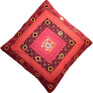 May include: Square decorative pillow with a patchwork design in shades of red, burgundy, and pink. The pillow features embroidered details, small mirrors, and a central square with a detailed pattern. The pillow is a square shape.
