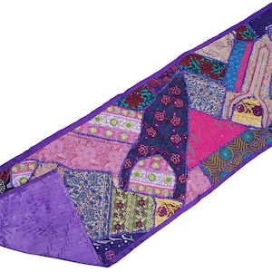 Può includere: Un runner da tavola rettangolare caratterizzato da un design patchwork di vari tessuti nei toni del viola, rosa, blu e verde. L'articolo è bordato da un tessuto viola a tinta unita e presenta ricami complessi, motivi floreali e forme geometriche.