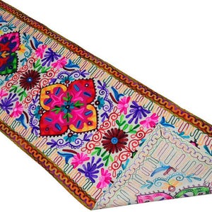 Puede incluir: Un camino de mesa rectangular con una base de color crema y bordados florales y geométricos multicolores. El diseño incluye grandes motivos de pétalos rosas, rojos y verdes, patrones de remolinos intrincados y un borde cosido marrón.