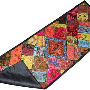 May include: A rectangular patchwork table runner with a black border and backing. The patchwork design features a variety of colourful squares with floral, elephant, and geometric patterns. The runner is approximately 152 cm long.