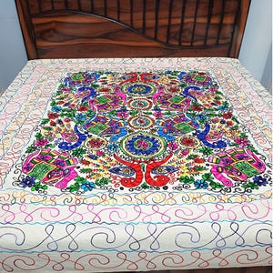 Puede incluir: Una colcha bordada y colorida con un diseño central de elefantes, pavos reales y motivos florales. La colcha es principalmente blanca con vibrantes acentos de rosa, azul, verde y amarillo. El borde tiene un patrón de remolino repetitivo.