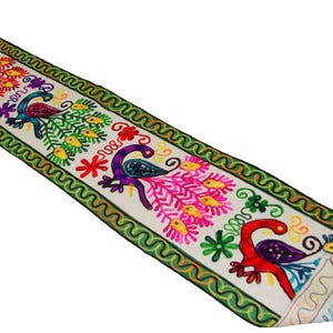 Puede incluir: Un camino de mesa largo y rectangular con una base de tela blanca y bordados multicolores vibrantes. El diseño muestra motivos de pavos reales repetidos en tonos morado, rojo y azul, acentuados con detalles florales amarillos y un borde verde ondulado.