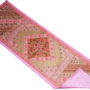 May include: A rectangular pink and beige embroidered table runner. The runner features a patchwork design with floral and geometric patterns. The edges are trimmed with a solid pink border. The runner is likely made of fabric.