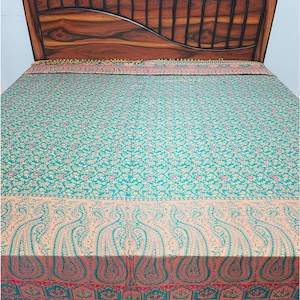 Op de afbeelding: Een rechthoekige sprei met een groenblauw en goudkleurig bloemmotief over het midden, een brede perzikkleurige rand met grote groenblauwe paisley-motieven en een onderkant afgewerkt met een rij donkerbruine decoratieve franjes.