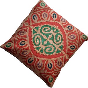 May include: Square cushion with a detailed, colourful design. The cushion has a central green swirl pattern within a red border, encircled by repeating oval shapes in various colours. The background is a light beige.