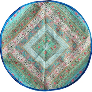 May include: A circular, quilted textile with a colourful design. The centre has a diamond pattern in teal and green, surrounded by floral and paisley motifs in pink, white, and gold. A bright blue border frames the piece.