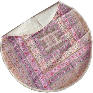 May include: A round, pink and cream-coloured decorative mat with an embroidered floral design. The mat features a central square pattern surrounded by striped panels with floral and leaf motifs. The back is a solid cream colour.