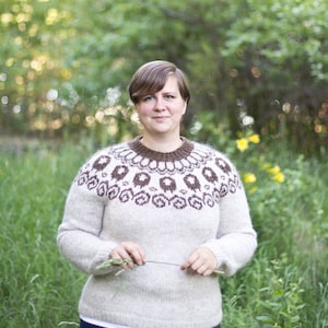 May include: A woman wearing a white and brown patterned sweater with a brown neckline. She is standing in a field of green grass and yellow flowers.