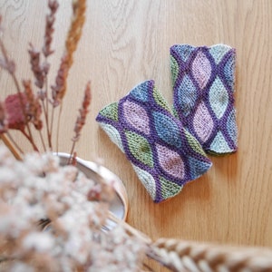 May include: A pair of knitted fingerless gloves with a geometric pattern in shades of green, purple, and blue. The gloves are laid out on a wooden surface.