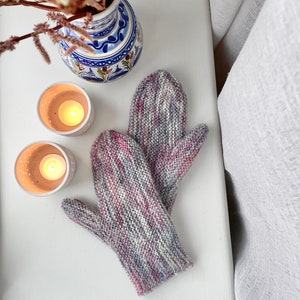 May include: A pair of knitted mittens in a mix of pink, purple, and gray. The mittens are on a white surface with two white candle holders and a vase of dried flowers.