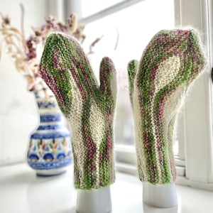 Puede incluir: Un par de mitones de punto de rayas verdes, blancas y rosas sobre un fondo blanco. Los mitones se muestran en soportes blancos.