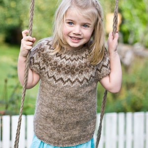 May include: A young girl with blonde hair is smiling while holding onto a rope swing. She is wearing a brown and white knitted sweater with a blue skirt.
