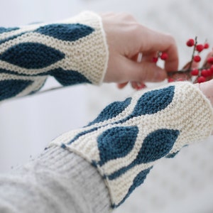 Puede incluir: Un par de calientamanos de punto con un patrón de hojas blancas y azul verdoso. Los calientamanos están hechos con un hilo suave y son perfectos para mantener las manos calientes en invierno.