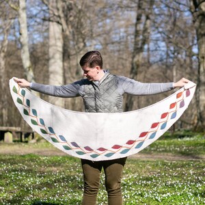 以下が含まれることがあります： グレーのベストと緑色のズボンを着用した人が、縁にカラフルな葉のデザインが施された白いクロシェのショールを持っています。