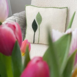 Può includere: Un cuscino quadrato decorativo con sfondo color crema e bordo verde. Il cuscino presenta un disegno di due foglie stilizzate in tonalità di verde e nero. L'immagine include anche tulipani rosa.