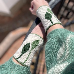 Puede incluir: Calentadores de muñeca de punto color crema con un diseño de hojas verdes. Los calentadores de muñeca tienen un borde verde y se usan con un suéter verde. El diseño de hojas es en tonos verdes.