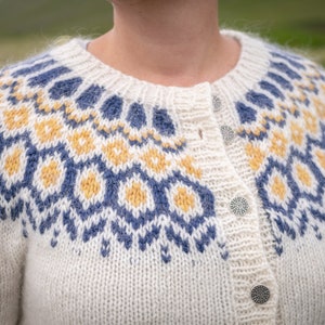 May include: A white knitted cardigan with a blue and yellow geometric pattern. The cardigan has a button closure and a fuzzy texture.