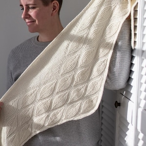 May include: Cream-colored knitted shawl with a diamond pattern. The shawl is held up, showcasing the intricate texture and design. The soft, neutral color and detailed knitwork suggest a cozy, handmade accessory.