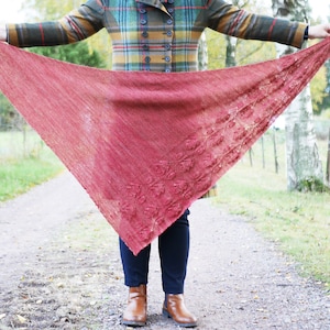 May include: A red knitted shawl with a delicate leaf pattern, held by a person wearing a plaid coat and jeans.