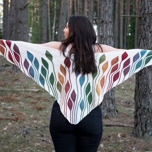 May include: A white knitted shawl with a rainbow pattern of leaves. The shawl is being held by a person in a forest setting.