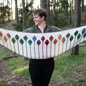 Op de afbeelding: Een witte gebreide sjaal met een herhaald patroon van kleurrijke, gestileerde bladvormen. De bladeren gaan over in tinten rood, oranje, geel, groen en blauw. De sjaal wordt voor een persoon in een bosrijke omgeving gehouden.