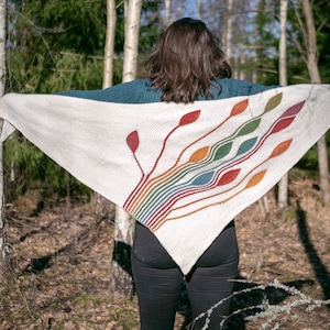 May include: A white knitted shawl with a rainbow and leaf design. The shawl is being worn by a person in a forest setting.
