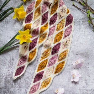 Könnte beinhalten: Ein weißer gestrickter Schal mit einem Rautenmuster in Burgunder- und Senfgelbtönen. Der Schal liegt auf einer grauen Oberfläche mit gelben Narzissen und rosa Orchideen.