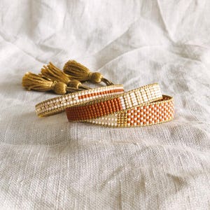 May include: Three beaded bracelets in the image. The bracelets are in shades of white, gold, and orange. The bracelets are made of small beads woven together. Gold tassels are in the background. The bracelets are on a white, textured surface.