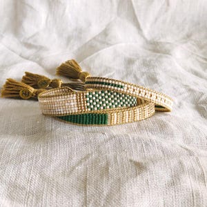 May include: Three beaded bracelets in cream, green, and gold are stacked on a white linen surface. The bracelets feature intricate woven patterns and gold accents. Tassels with gold-coloured charms are in the background.