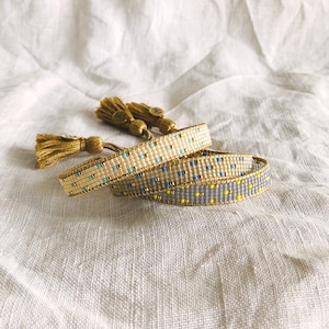 May include: Two beaded bracelets with gold detailing and tassels. One bracelet is beige with blue and gold beads, the other is grey with yellow and gold beads. The tassels are gold.