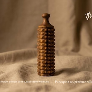 May include: A cylindrical brown wooden acupressure roller featuring a textured surface with numerous small, pyramid-shaped spikes. The top has a rounded knob handle. Text on the image reads: Handmade; ethical and sustainable wellness Porcupine acupressure roller.