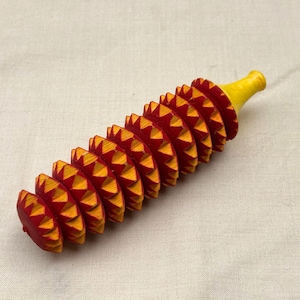 May include: A cylindrical wooden massage tool featuring a series of stacked, gear-shaped discs painted in alternating red and yellow. The central shaft is topped with a smooth, solid yellow handle, and the edges of each disc are serrated.