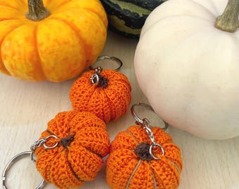 May include: Three crocheted orange pumpkin keychains with brown stems and silver key rings. The keychains are next to a white pumpkin and a green and brown gourd.