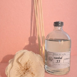 Op de afbeelding: Een heldere glazen diffuserfles met een zwarte dop, gevuld met vloeistof, en een label met de tekst "santal 33". Een bundel houten rietjes en een stoffen bloem staan naast de fles. De achtergrond is effen roze.