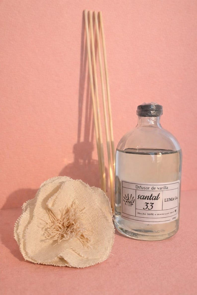 Puede incluir: Una botella difusora de vidrio transparente con tapa negra, llena de l&iacute;quido, y una etiqueta que dice "santal 33". Un manojo de ca&ntilde;as de madera y una flor de tela est&aacute;n junto a la botella. El fondo es rosa liso.