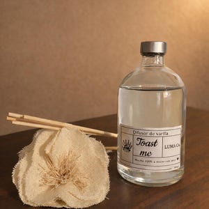 Puede incluir: Una botella difusora de vidrio transparente con tapa plateada, llena de l&iacute;quido. La etiqueta dice "Toast me" y "Difusor de varilla". Una flor de tela beige y palitos de madera est&aacute;n junto a la botella. Para fragancia del hogar.
