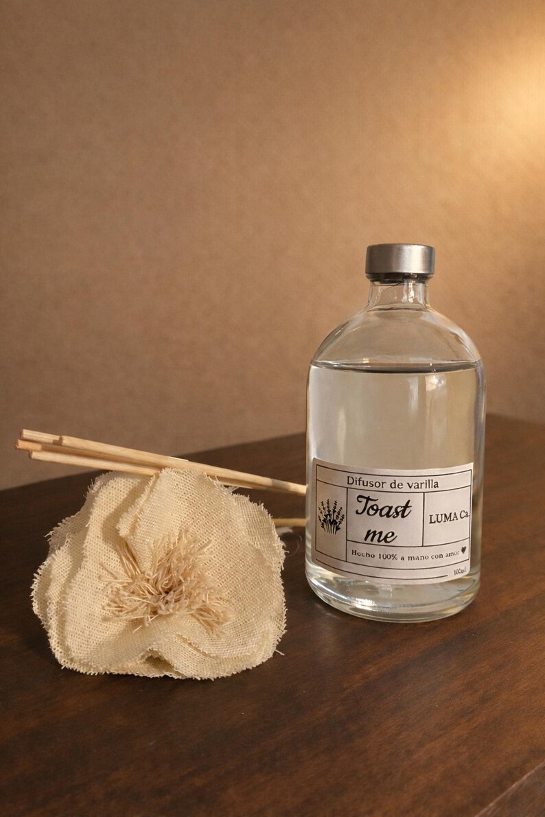 Puede incluir: Una botella difusora de vidrio transparente con tapa plateada, llena de l&iacute;quido. La etiqueta dice "Toast me" y "Difusor de varilla". Una flor de tela beige y palitos de madera est&aacute;n junto a la botella. Para fragancia del hogar.