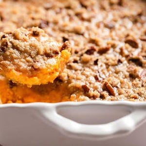 May include: A close-up of a baked sweet potato casserole in a white baking dish. The casserole has a golden-brown, crumbly topping with visible pecans. A spoonful of the casserole is being lifted from the dish, showing the orange filling.