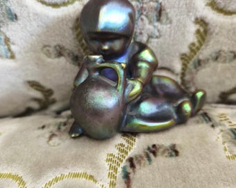 Peut inclure: Petite figurine irisée représentant un enfant assis tenant une cruche. La figurine présente une finition nacrée avec des teintes de bleu, de vert et de violet. L'enfant porte un bonnet et des vêtements, et la cruche a une anse.