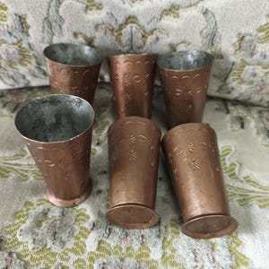 May include: Six copper-coloured metal cups with a decorative floral pattern. The cups have a flared shape and a silver interior. The cups are arranged on a patterned fabric.