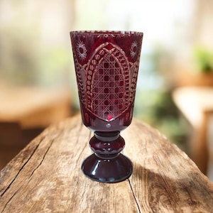 May include: A ruby red, cut crystal goblet with intricate geometric patterns. The goblet features a detailed design on the bowl and a rounded stem. The glass is approximately 18 cm tall, perfect for serving beverages.