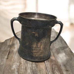 Può includere: Un boccale d'argento ossidato con due manici, appoggiato su una superficie di legno stagionato. Il boccale ha un bordo ampio e leggermente angolato e un corpo cilindrico. Il metallo mostra segni di età e usura.