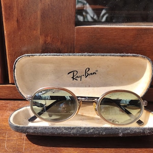 Peut inclure: Lunettes de soleil ovales Ray-Ban avec des montures métalliques fines et des verres teintés foncés, reposant dans un étui rigide. L'intérieur de l'étui présente une doublure de couleur crème avec le logo Ray-Ban imprimé en noir.