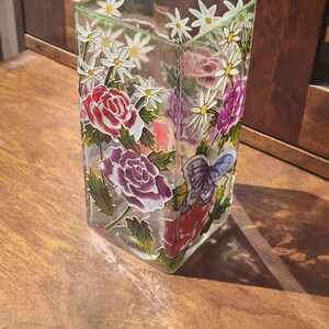 May include: A clear, square glass vase featuring hand-painted floral designs including red and purple roses, white daisies, and a blue butterfly. The painted motifs are outlined in black, creating a stained-glass effect on the transparent glass surface.
