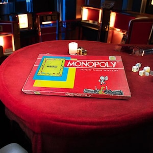 May include: A vintage Monopoly board game on a red table. The game box is red with the Monopoly logo and game board graphics. Dice and a candle are also on the table. The background includes retro-style furniture.