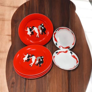 Può includere: Un set di piatti in ceramica rossa con un design di figure danzanti in bianco e nero. Sono presenti anche due piatti bianchi più piccoli con motivi astratti rossi e neri. I piatti sono disposti su una superficie di legno scuro.
