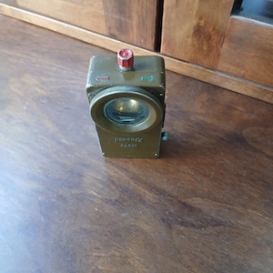 May include: A rectangular olive green metal flashlight with a circular lens, a red push-button switch on top, and directional arrows in red and green. The device features the embossed text "PERDIX" and "No. 620" on the front face below the lens.
