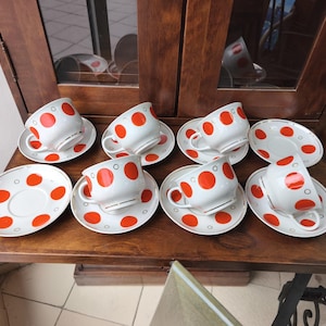 May include: A set of eight white ceramic teacups and saucers with a red polka dot pattern. The cups have a curved handle and the saucers are round. The set is displayed on a wooden surface.